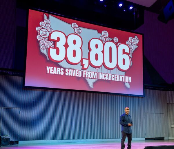 Raj Jayadev of Silicon Valley De-Bug speaks on stage while a red image displayed on the screen behind him reads "38,806 year saved from incarceration"