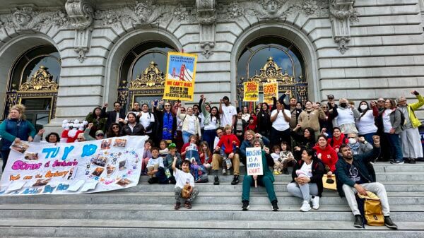 Group of people of various ages hold signs and lifted fists on steps of city hall