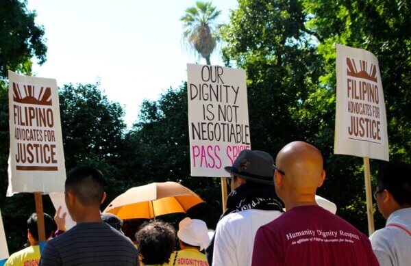Community members gather at an outdoor rally holding signs from Filipino Advocates for Justice and a central message reading “Our dignity is not negotiable,” highlighting collective advocacy and community support.