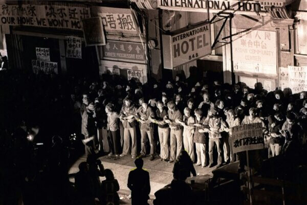 Black-and-white photo of a large crowd gathered outside the International Hotel, where a line of people link arms across the entrance as others stand behind them; protest signs and storefronts with Chinese characters are visible above.