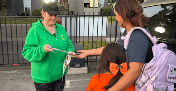 A community outreach worker in a bright green sweatshirt hands a flyer to an adult standing with a child in a residential parking lot at dusk. Apartment buildings, trees, and a parked vehicle are visible in the background, while the adult rests an arm around the child during the exchange.
